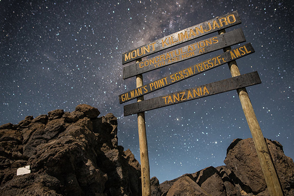 Gilman’s Point Mount Kilimanjaro summit sign under starry night sky showing elevation and rocky volcanic landscape.